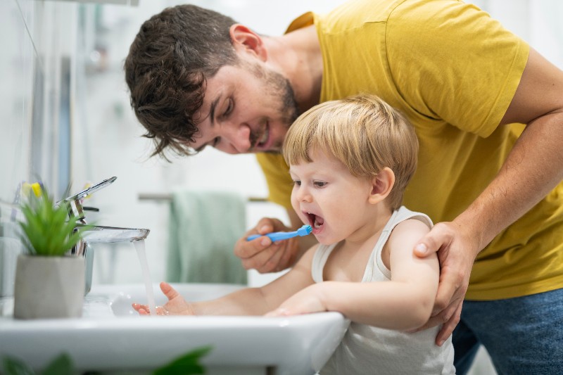 A man brushing a toddler boy’s teeth in a sink by Villanova Dental Studio