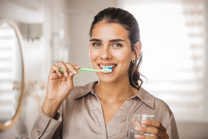 A woman brushing her teeth and holding a glass of water by Villanova Dental Studio