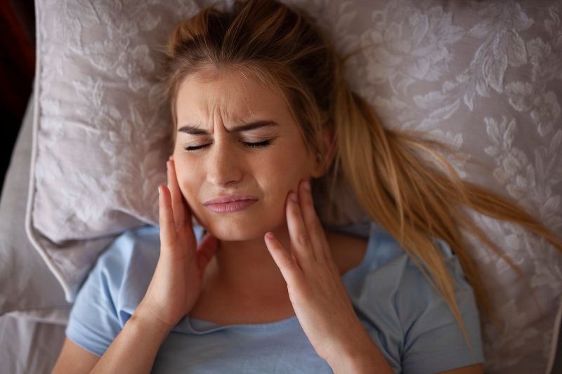 Woman experiencing jaw pain and facial discomfort from teeth grinding or TMJ issues while lying in bed in Kanata and Stittsville, ON